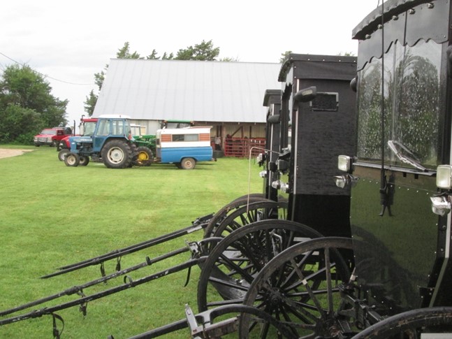 Amish buggies at an auction in Kansas. In the background are Amish “pickup trucks” – tractors with a pickup truck bed trailer for hauling family and supplies.